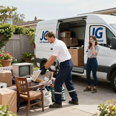 Man loading boxes into a moving van, woman on phone nearby. House in background.