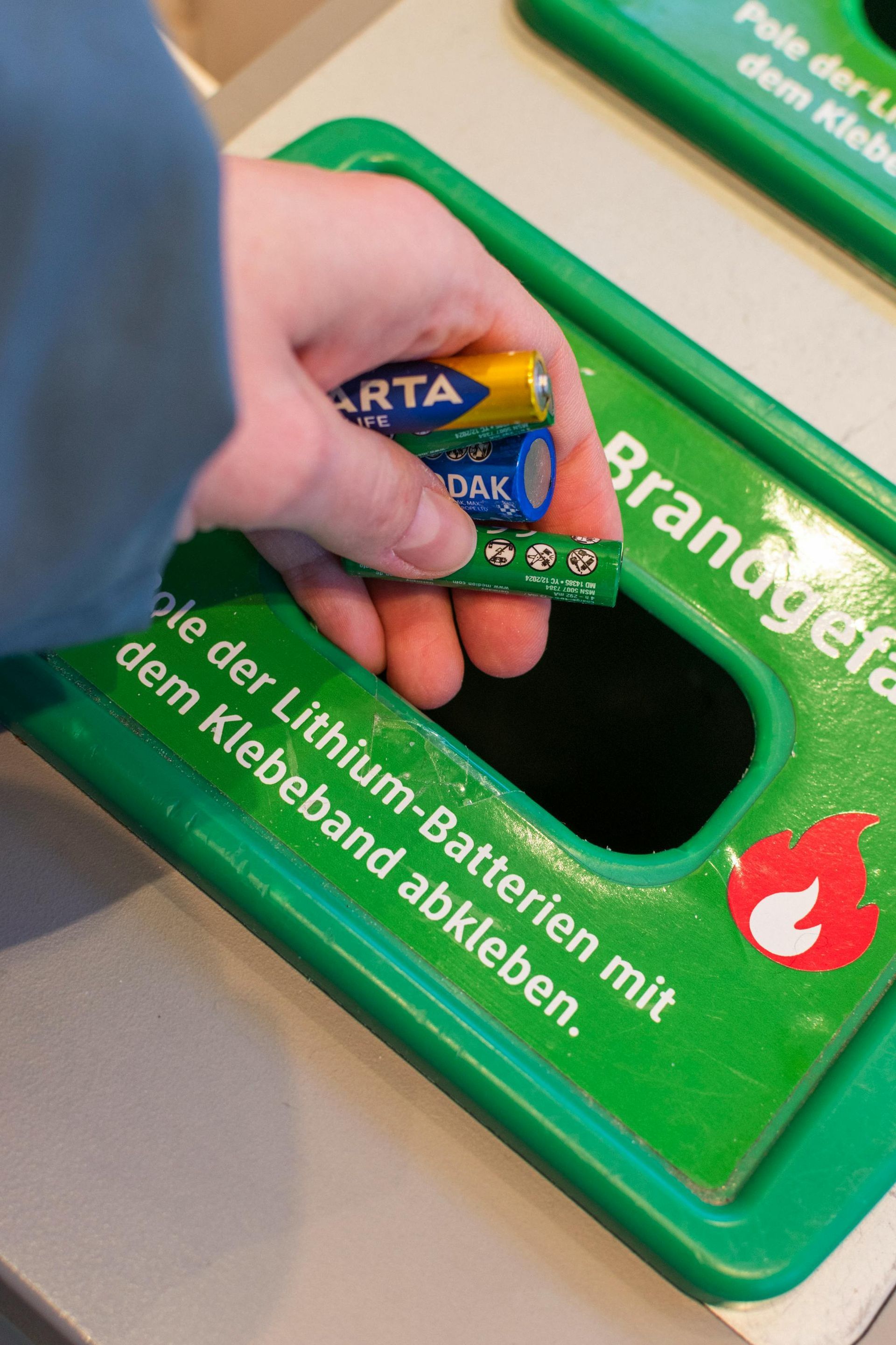 Hand placing batteries and a circuit board into a green recycling bin. German text on bin.