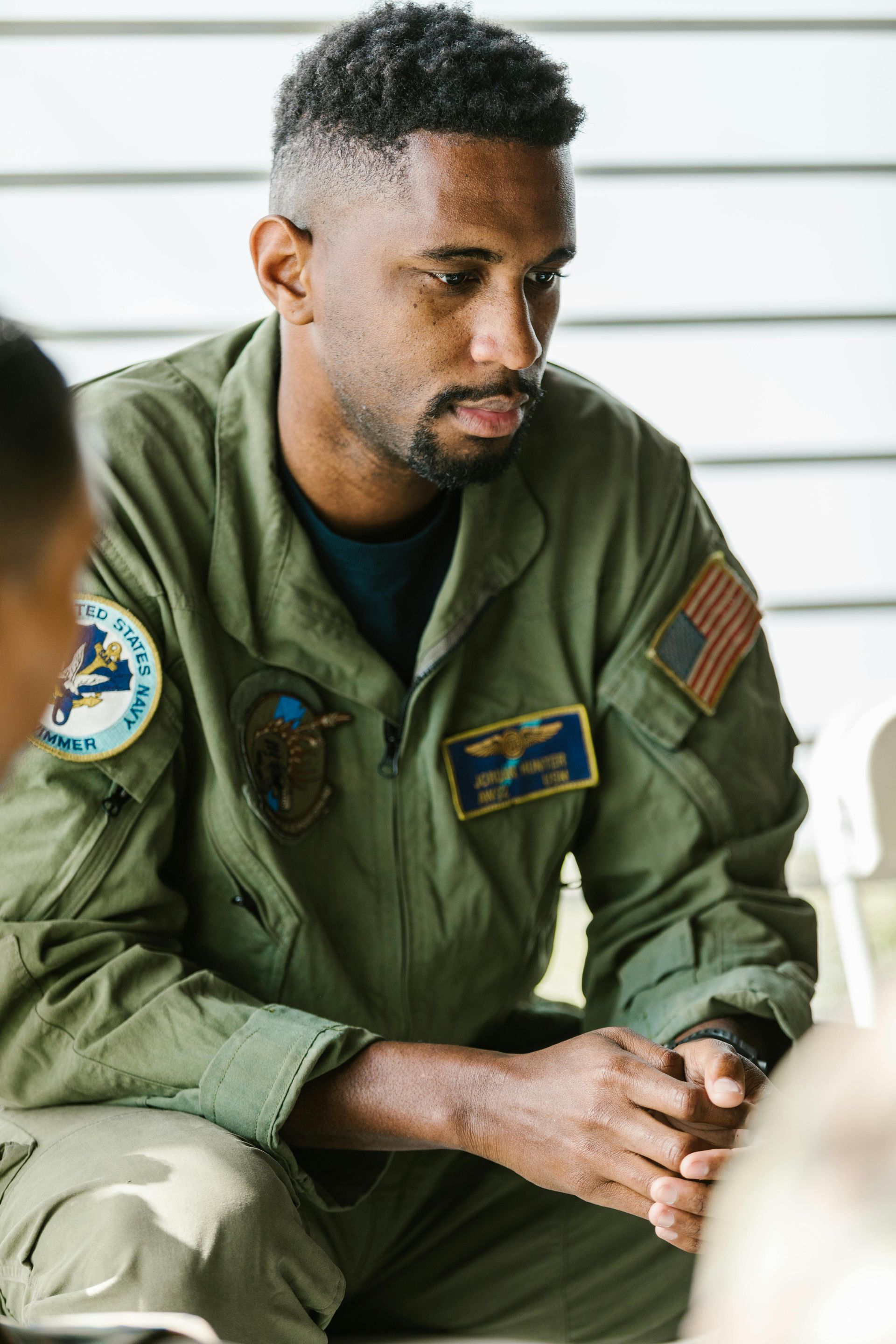 Man in olive-green flight suit, seated, looking down thoughtfully, possibly in a group setting outdoors.