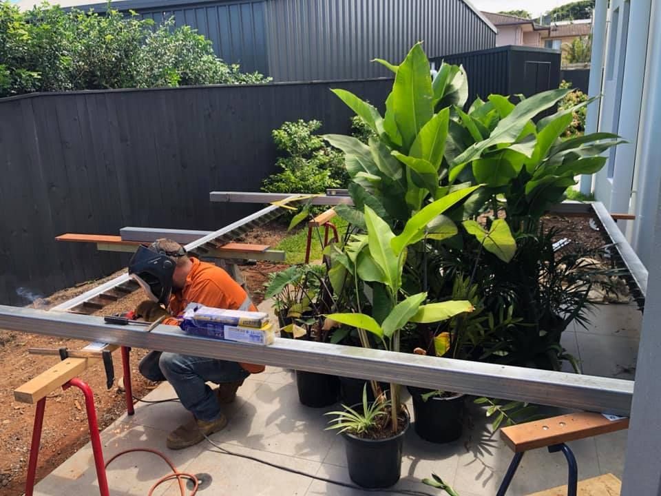 A Man is Welding a Metal Frame in a Backyard Next to Potted Plants — Jensen Welding & Fabrication in Chevallum, QLD