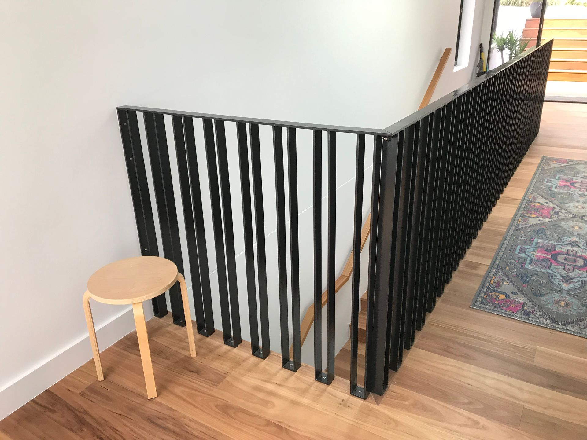 Black baby gate blocking a hallway, with a small wooden stool beside it on a hardwood floor. — Jensen Welding & Fabrication in Chevallum, QLD