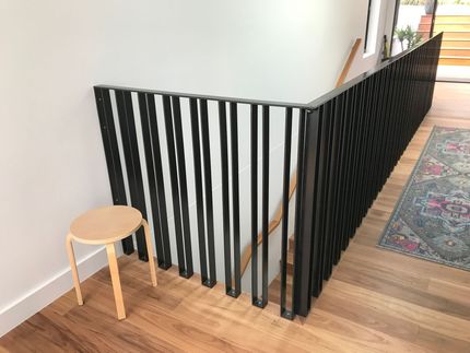 Black stair railing with vertical slats beside a small wooden stool in a bright hallway  — Jensen Welding & Fabrication in Chevallum, QLD