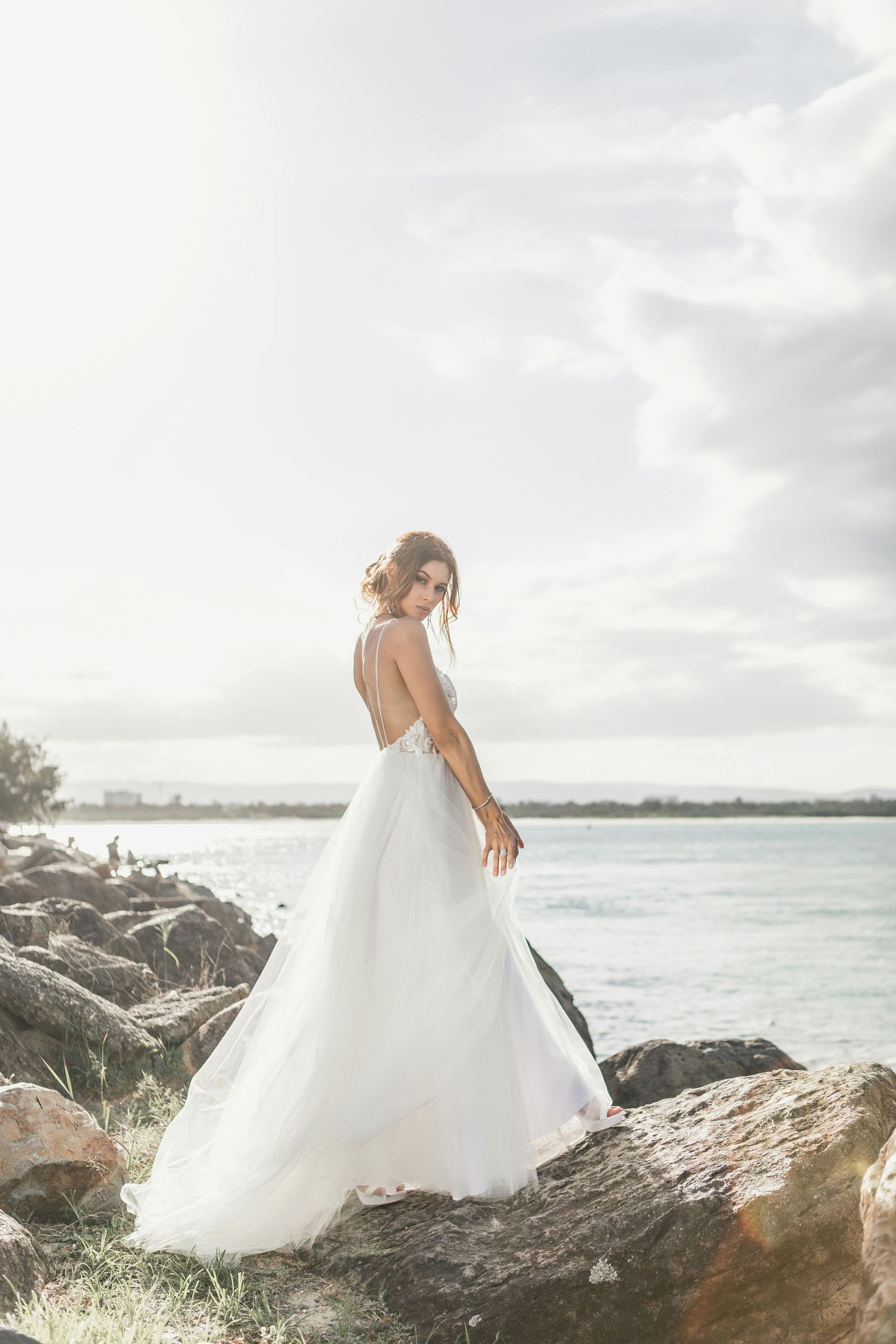 Une femme en robe de mariée se tient debout sur un rocher près de l'océan.