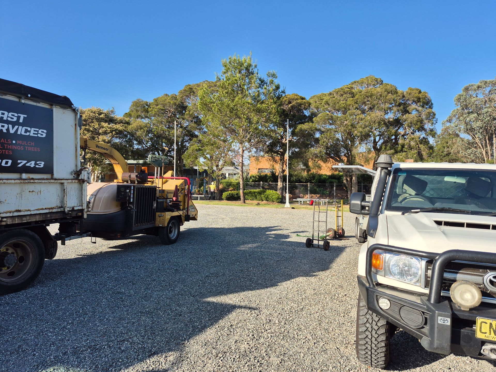 Truck, chipper, and white SUV parked on gravel, trees in the background, clear blue sky.
