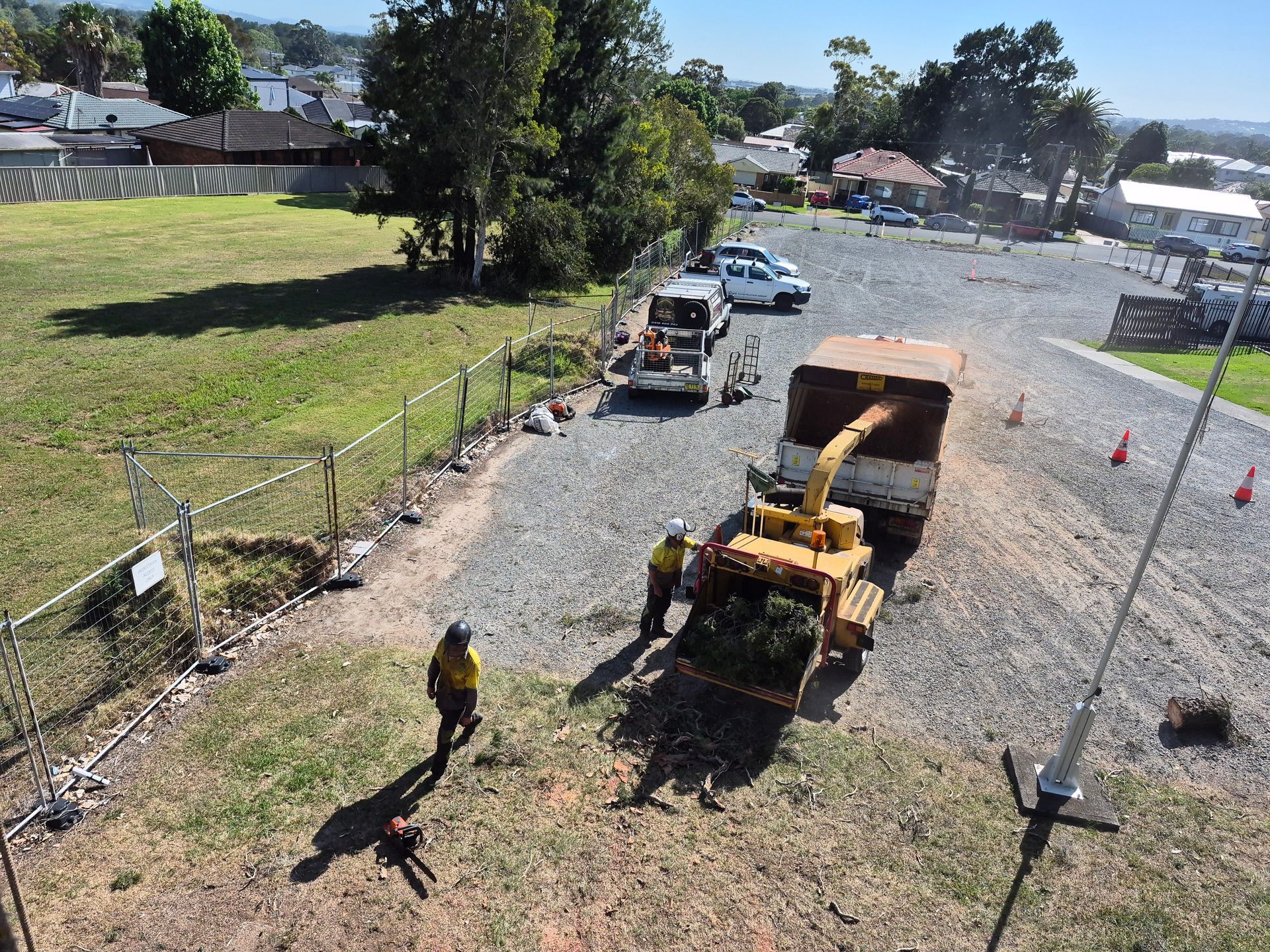 Workers using a wood chipper on a gravel lot; vehicles and houses in background.