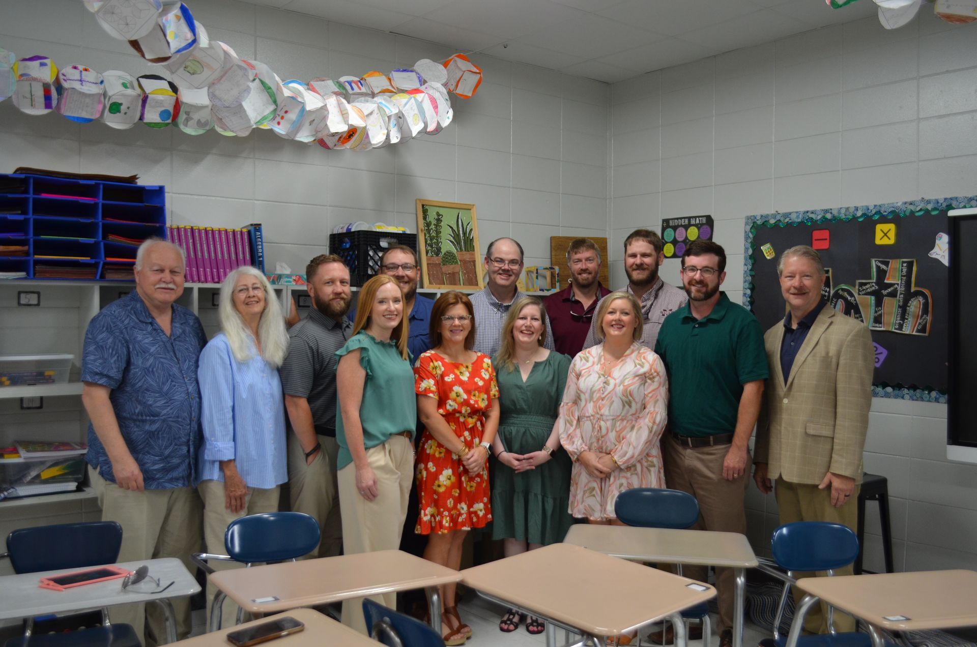 Group of adults in classroom, smiling at camera. Desks in front, artwork on wall.