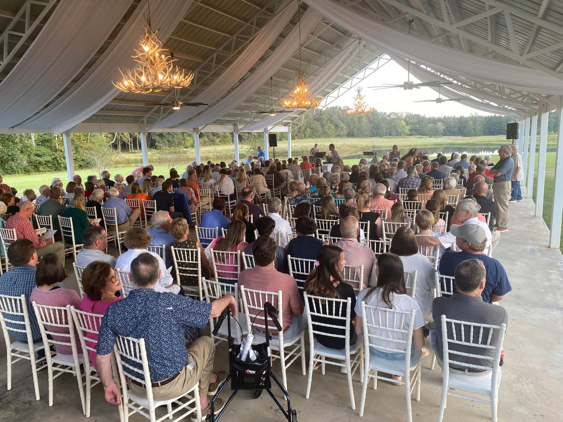 Large gathering of people seated under a white open-air tent, possibly an outdoor event or ceremony.