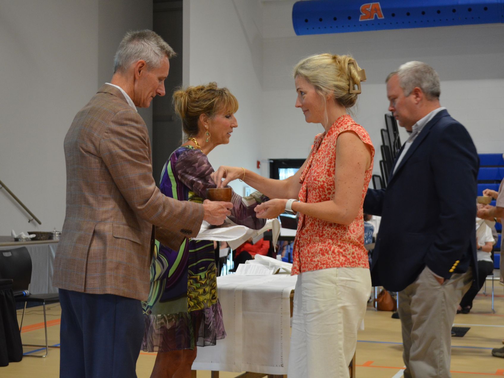 Four people at a table, two handing something to the other two. Indoors, brightly lit.