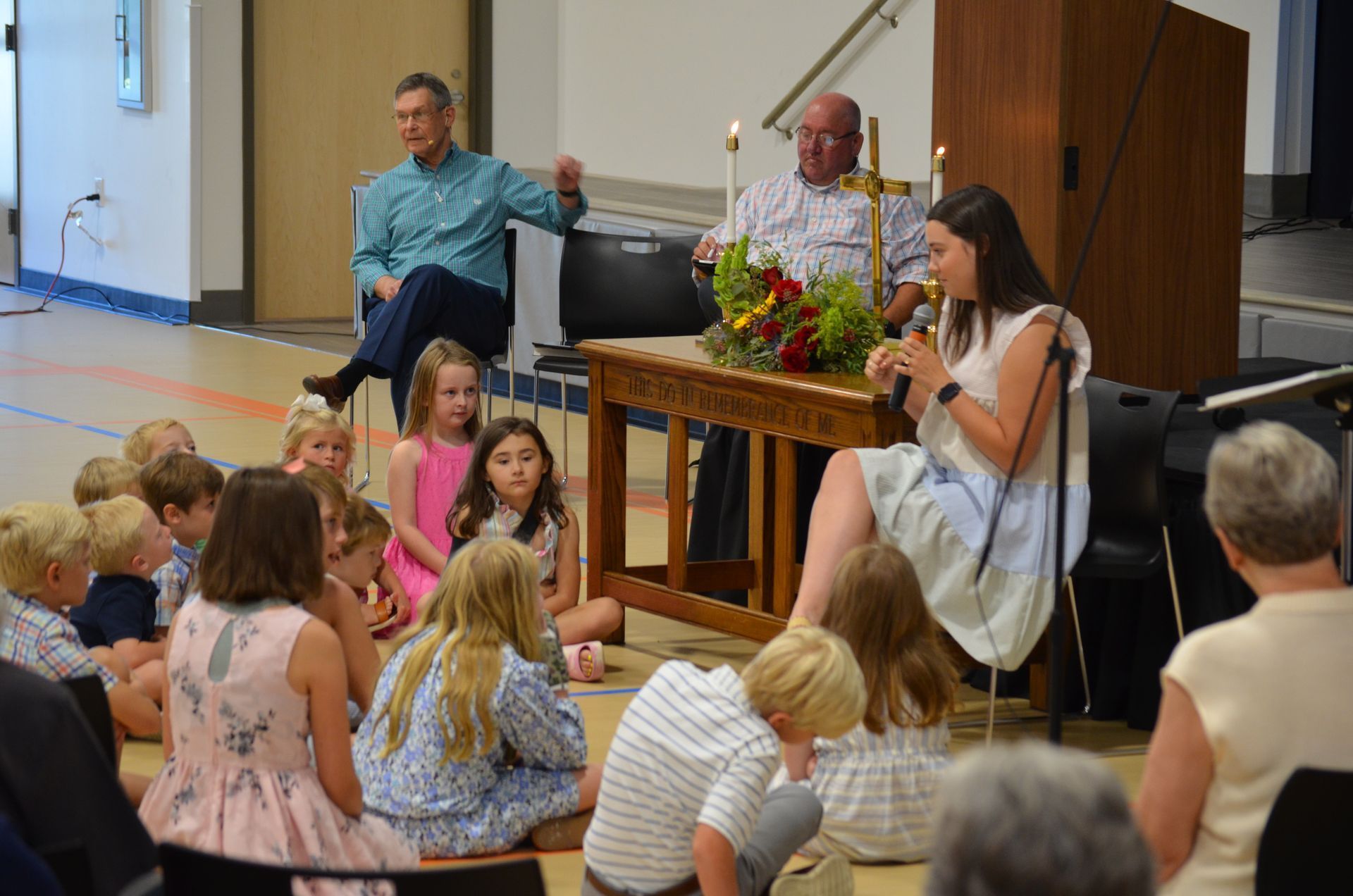 A woman speaks to children seated on floor; two men sit behind a table with flowers.