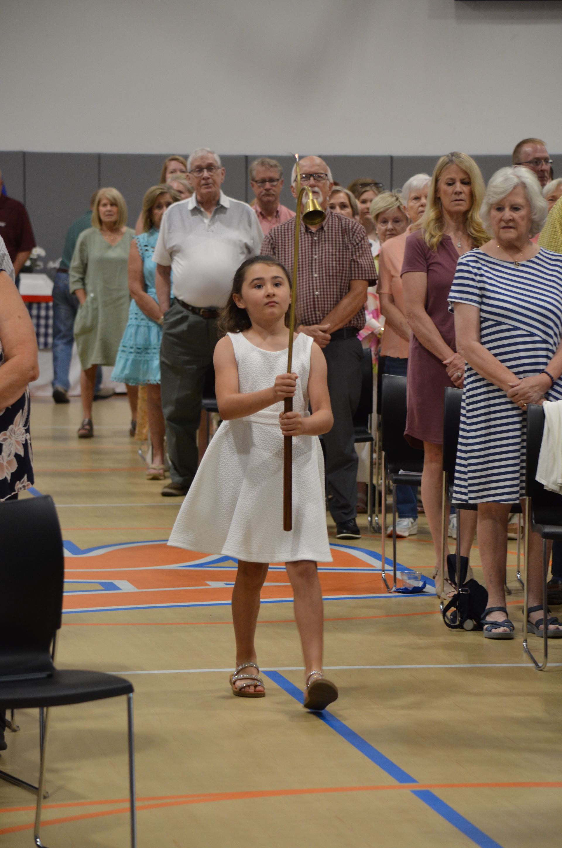 Girl in white dress holding bell, walking down aisle, people watching. Gymnasium.