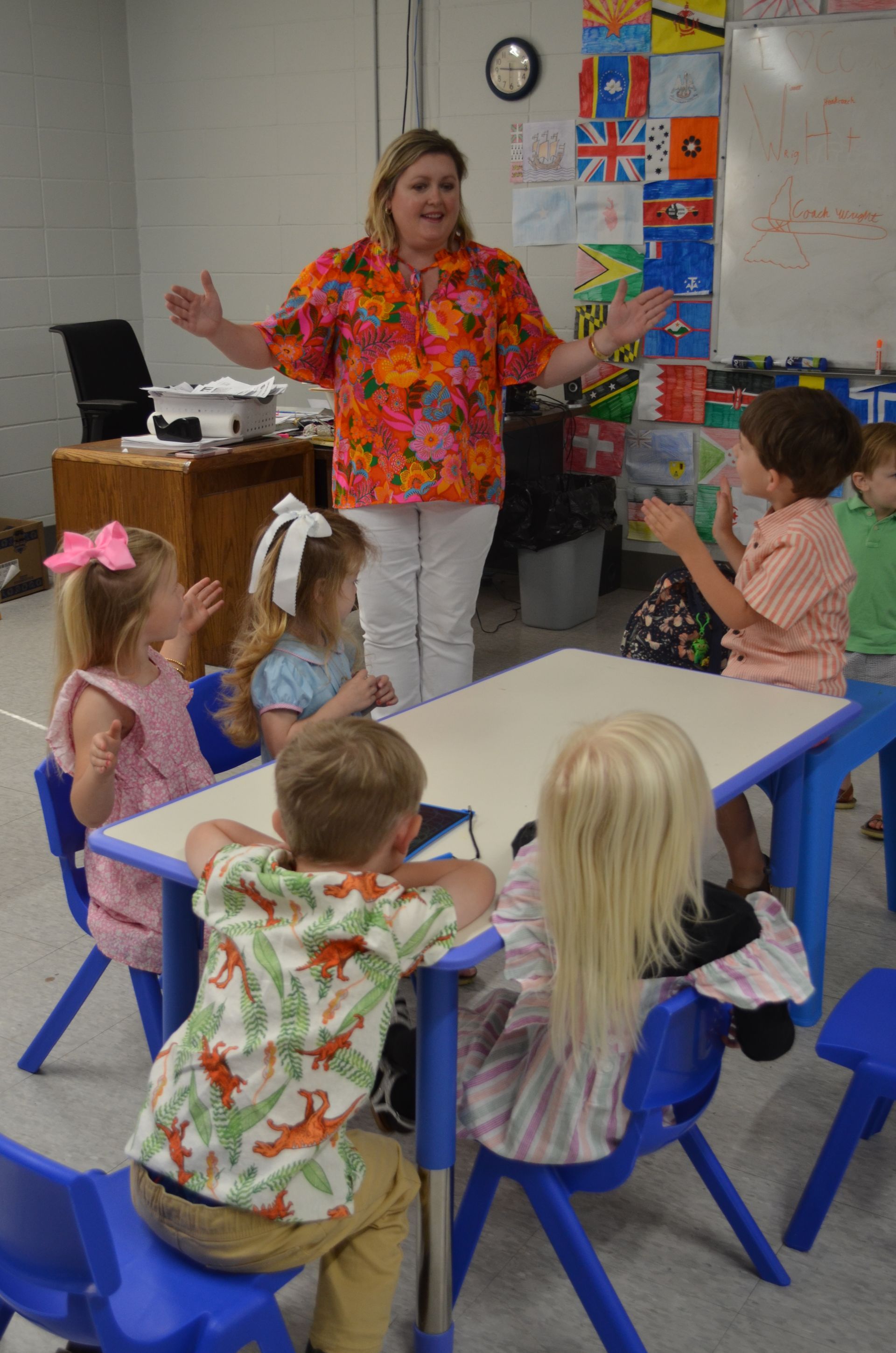 A woman is standing in front of a group of children sitting at tables.