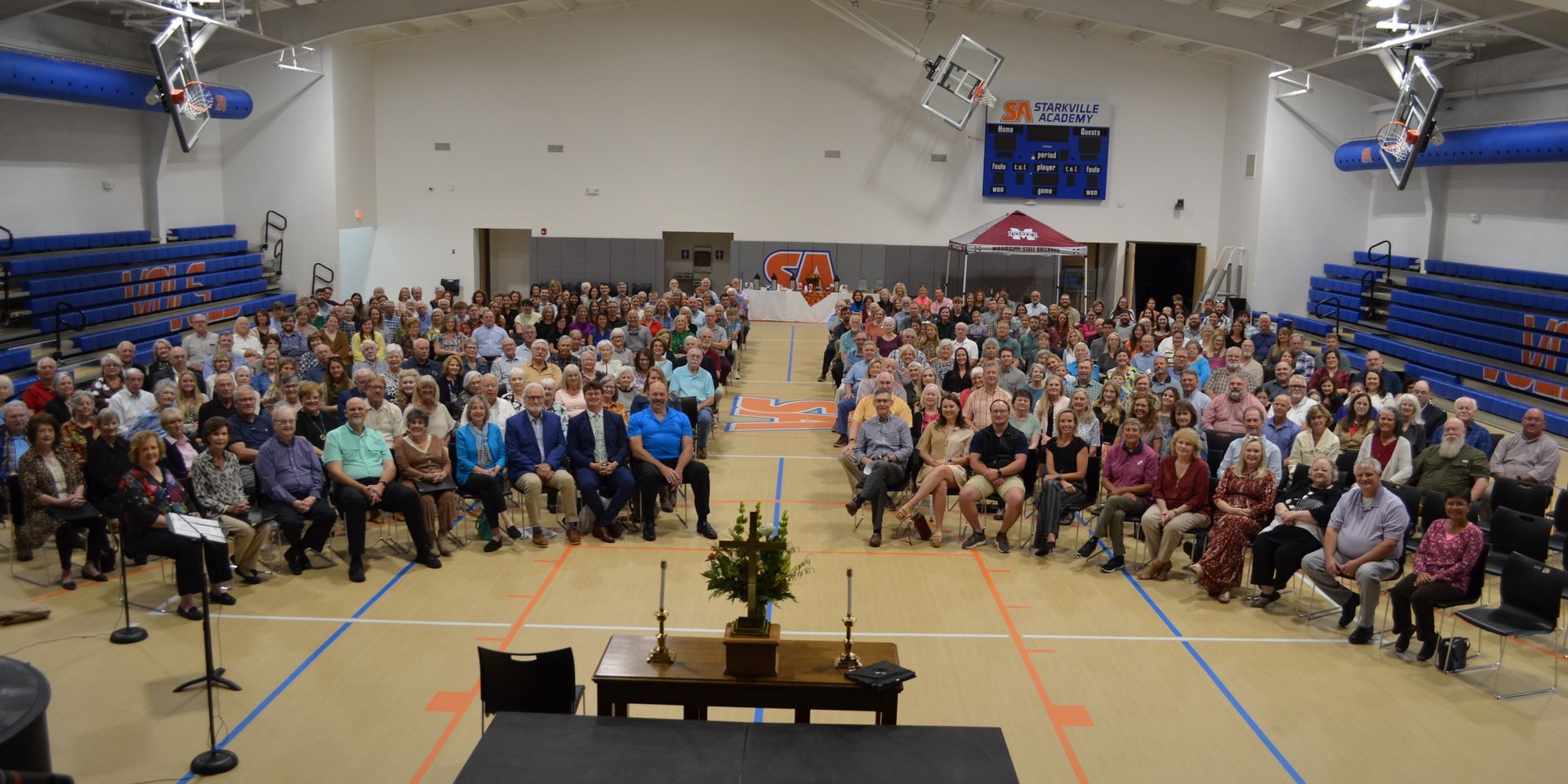 Large crowd seated in a gymnasium; central table with floral arrangement; basketball court markings.