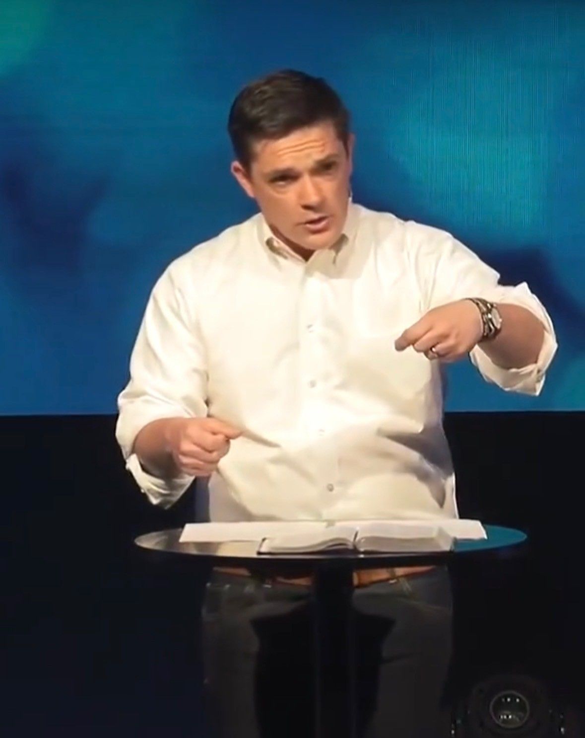 Man in white shirt gestures over open book at a podium with blue backdrop.