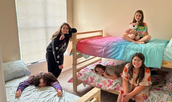 Four girls in a bedroom with bunk beds. One leans on a bed, three sit on or near beds, smiling.