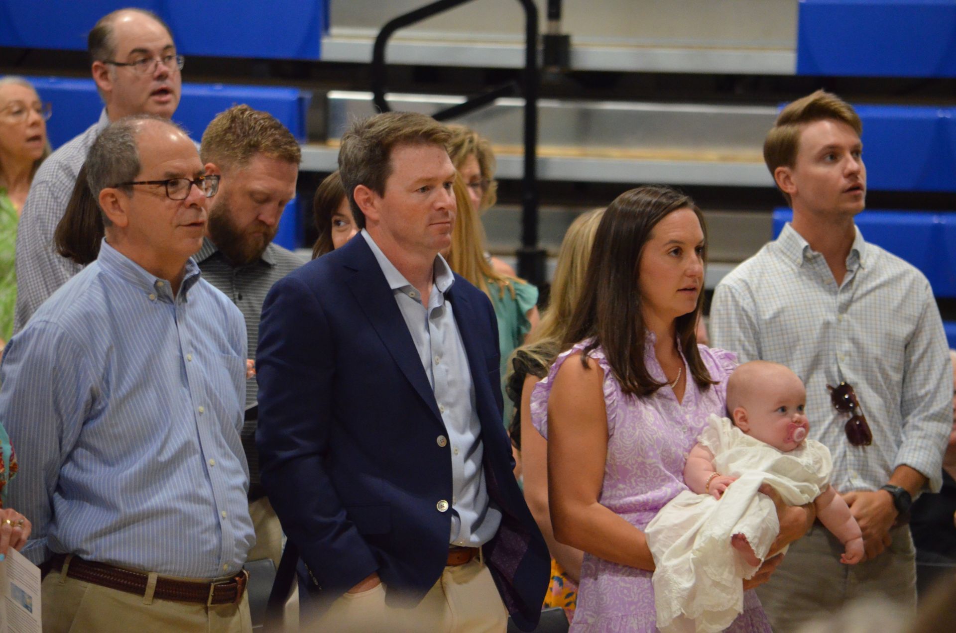 People in a gymnasium, woman holding baby, man in blazer, others with serious expressions.