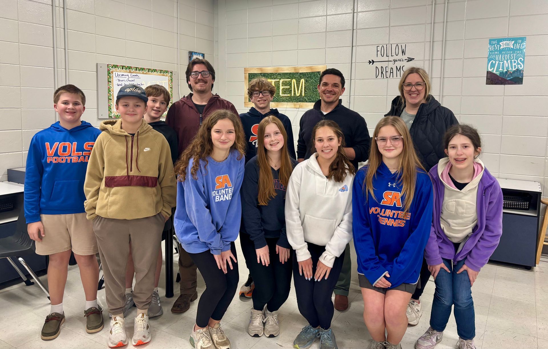 Group of students and adults posing in a classroom; smiles, casual attire, neutral background with posters.