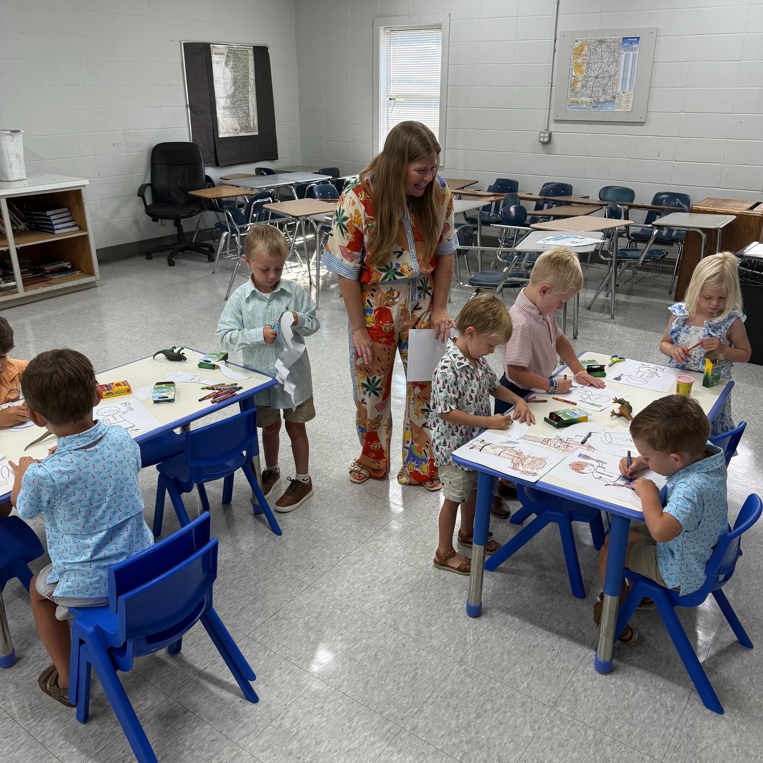 A teacher and children at tables in a classroom are doing an activity.