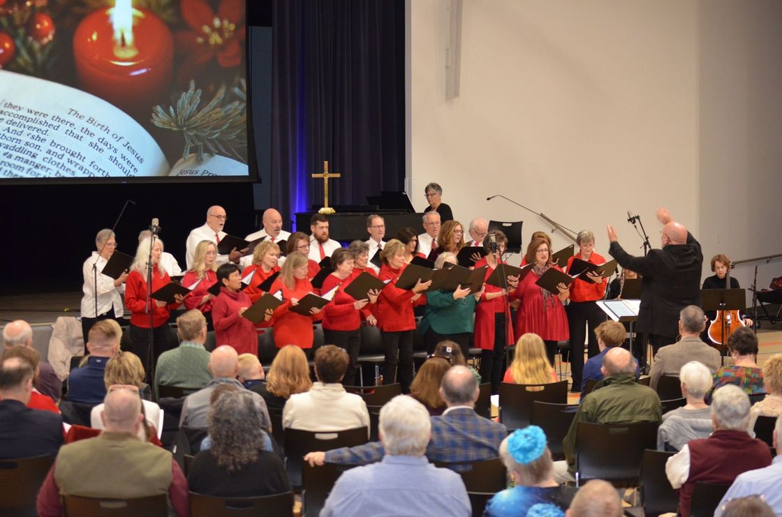 Choir performing on stage in front of an audience. Red and white outfits, conductor with raised arms, cross backdrop.