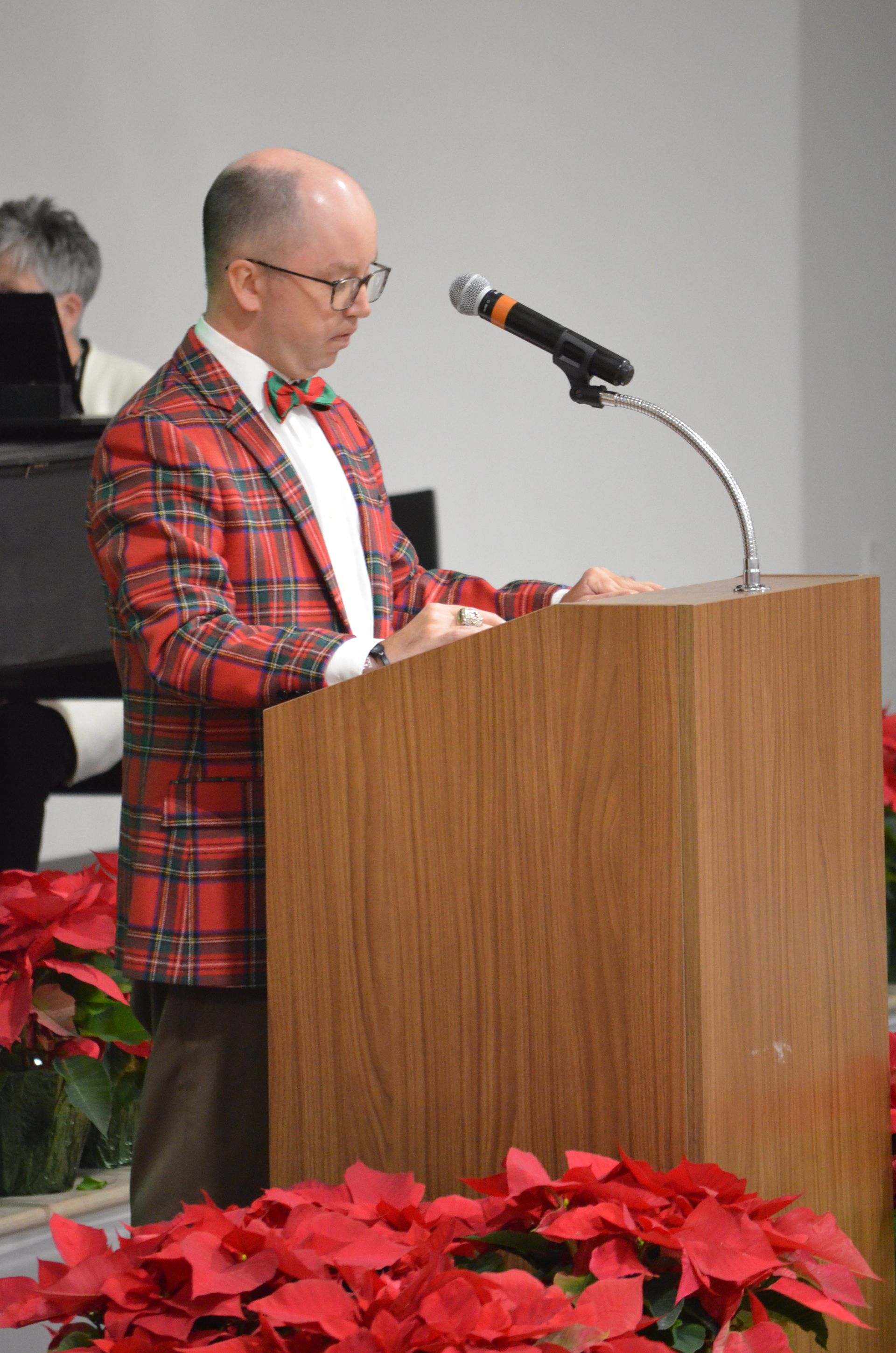 Man in plaid blazer speaks at a wooden podium, with poinsettias in the foreground.