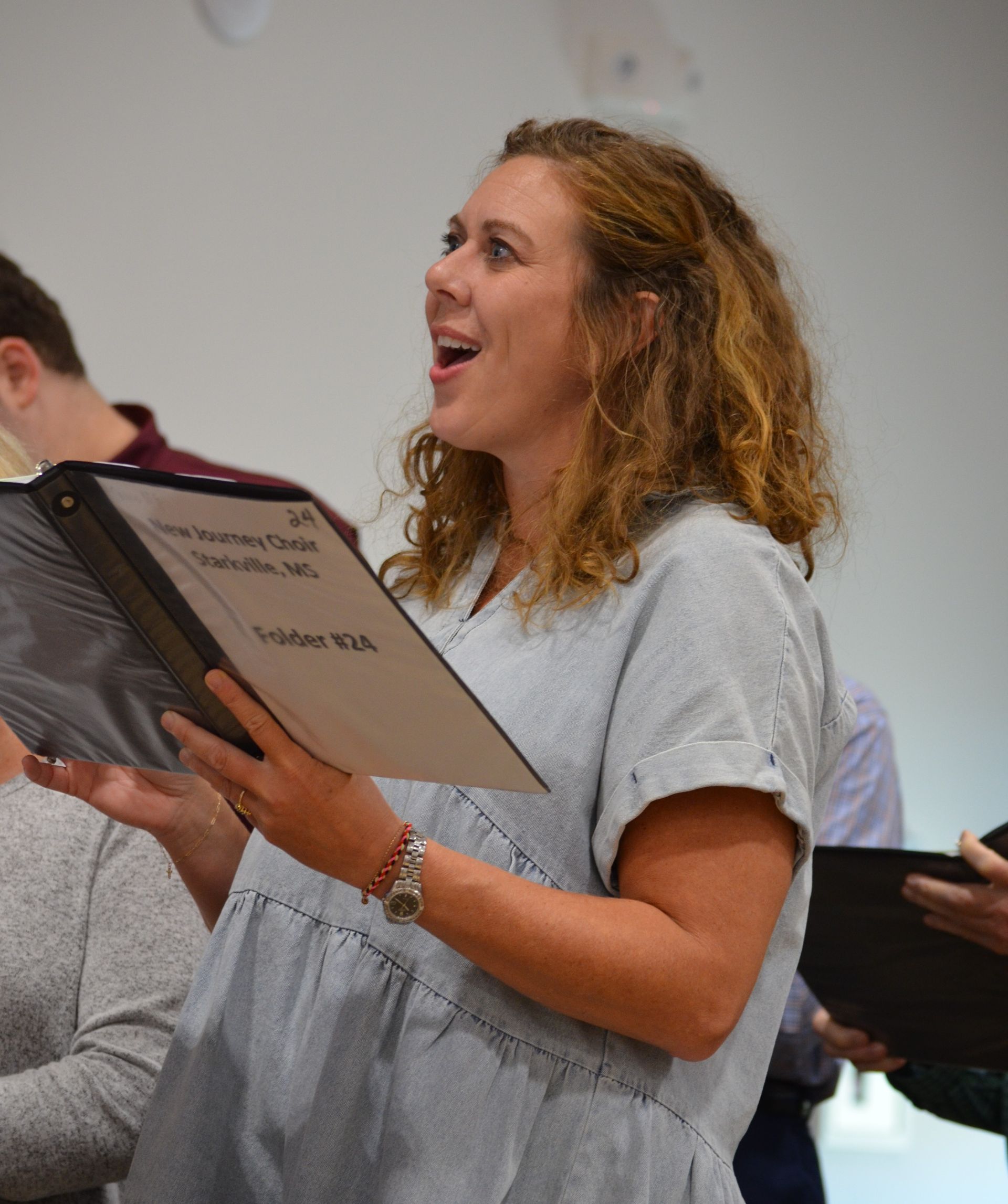 Woman singing from sheet music, wearing gray dress, mouth open, indoors.