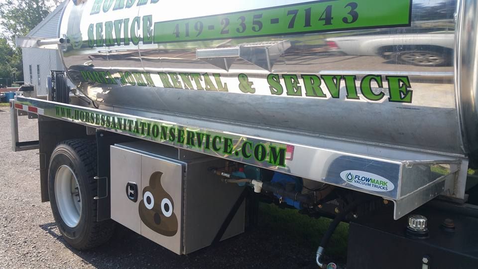 A close-up of a silver sanitation truck featuring a cartoon poop emoji decal on a side storage box.