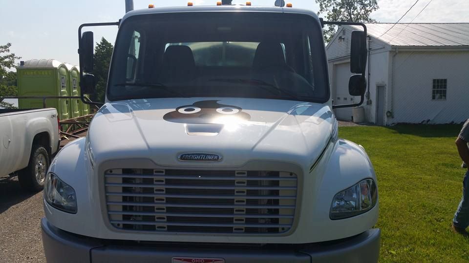 Front view of a white Freightliner truck with a poop emoji decal on the hood, parked near portable toilets and a building.