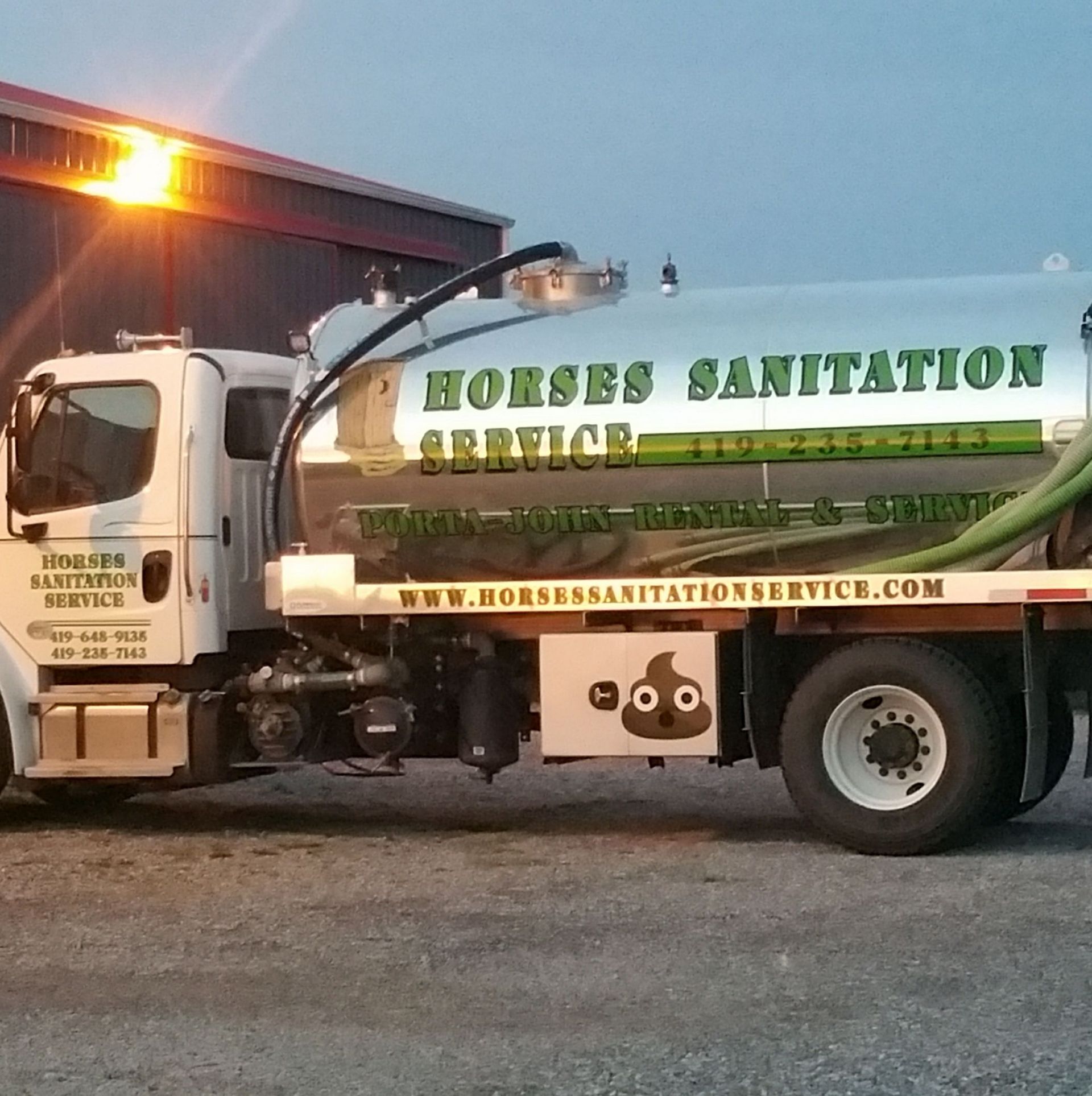 A white sanitation truck with a chrome tank, green text, and a poop emoji decal on the side, parked on gravel.