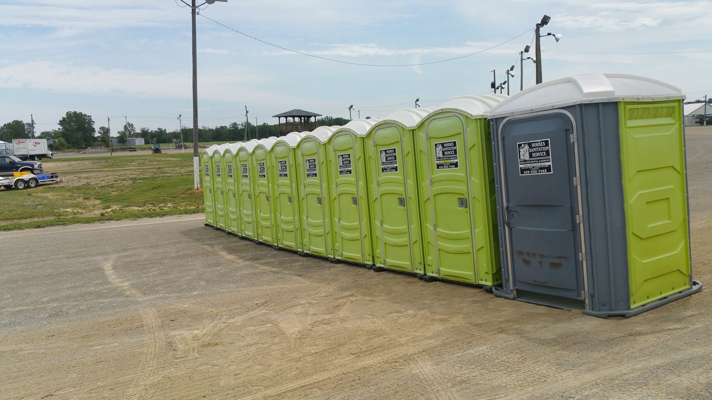 A long row of portable lime-green toilets set up on a gravel lot under a bright, overcast sky.