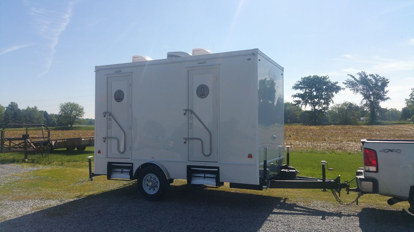 A white portable restroom trailer with two entrance doors and stairs, hitched to a pickup truck in a sunny field.