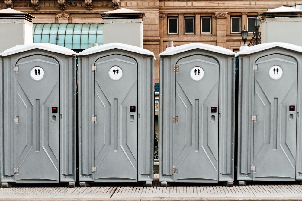 A row of four grey portable toilets stands side-by-side in front of a stone building.