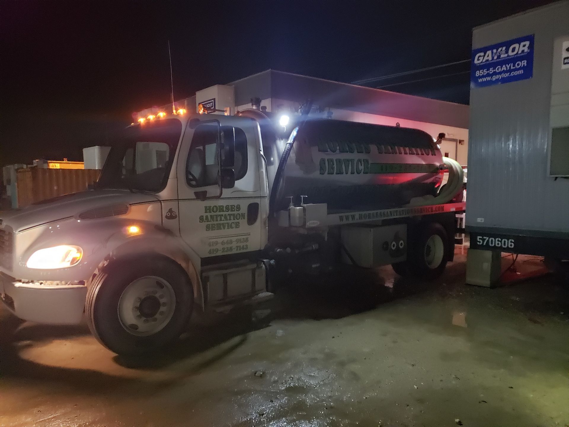 A white septic tank truck parked at night next to a portable office unit.