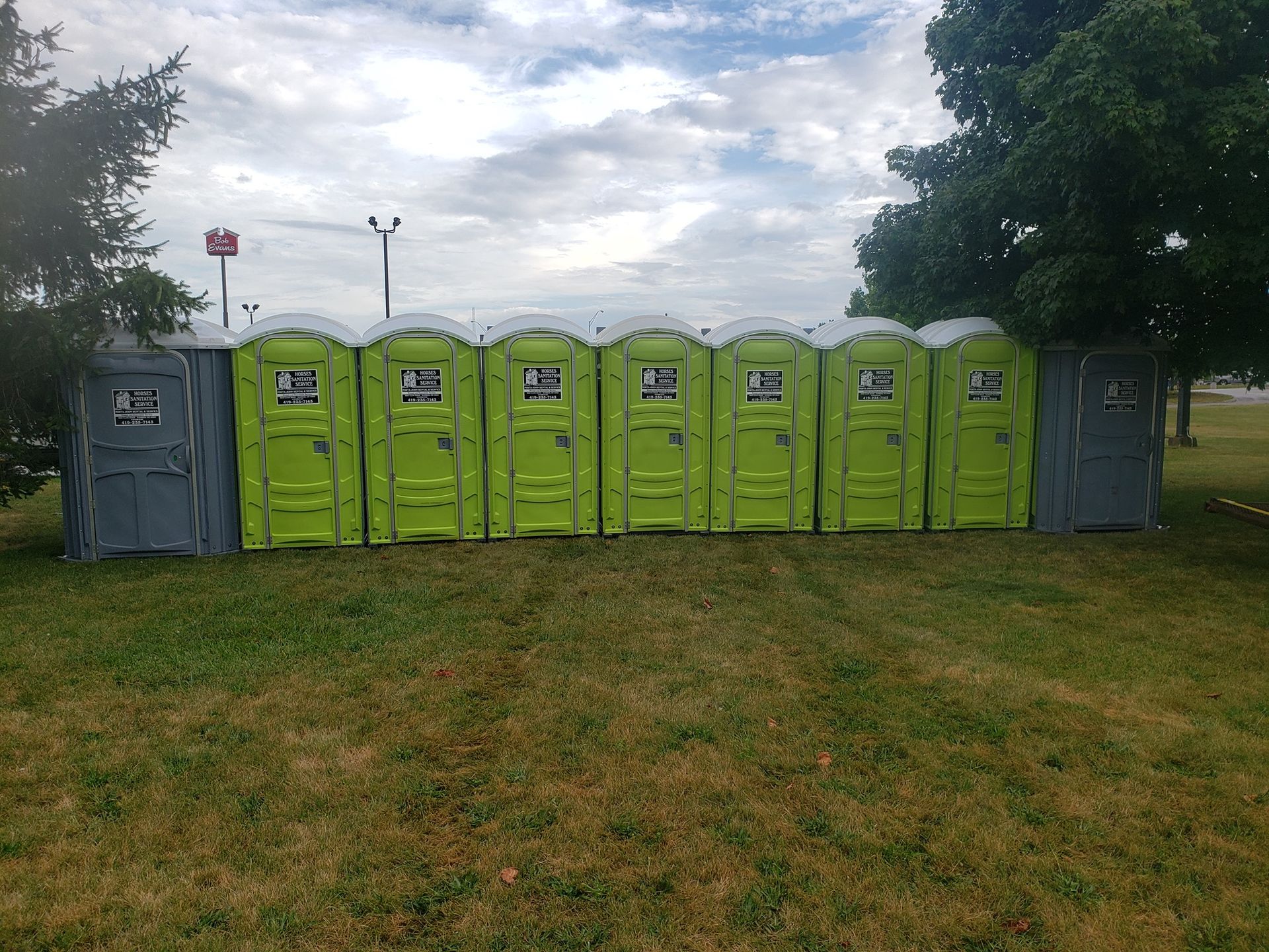 A row of eight portable toilets, six lime green and two gray, standing on a patch of grass under a cloudy sky.
