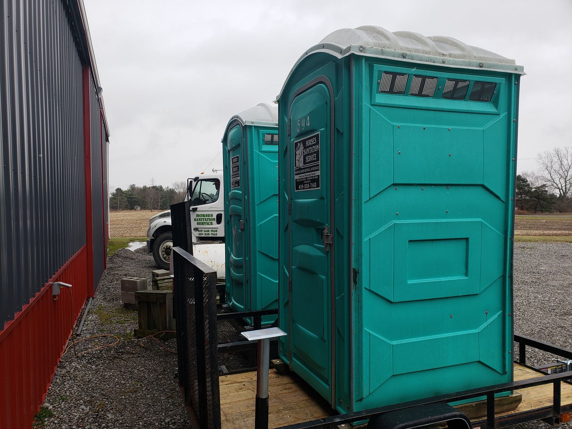 Two teal portable toilets on a trailer, positioned next to a red-and-gray building on a gravel lot.