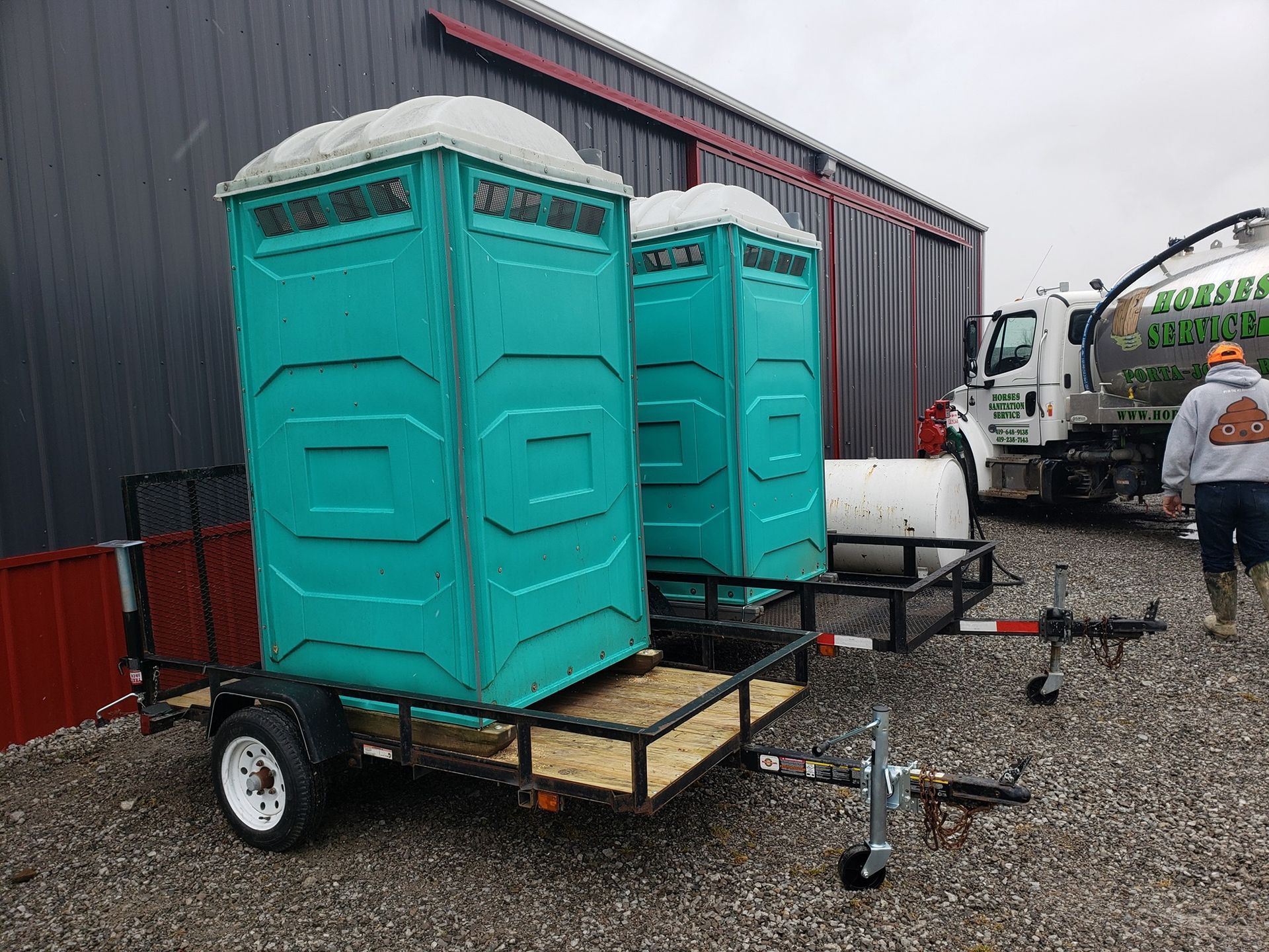 Two teal portable toilets on flatbed trailers parked on a gravel lot near a building and a service truck.