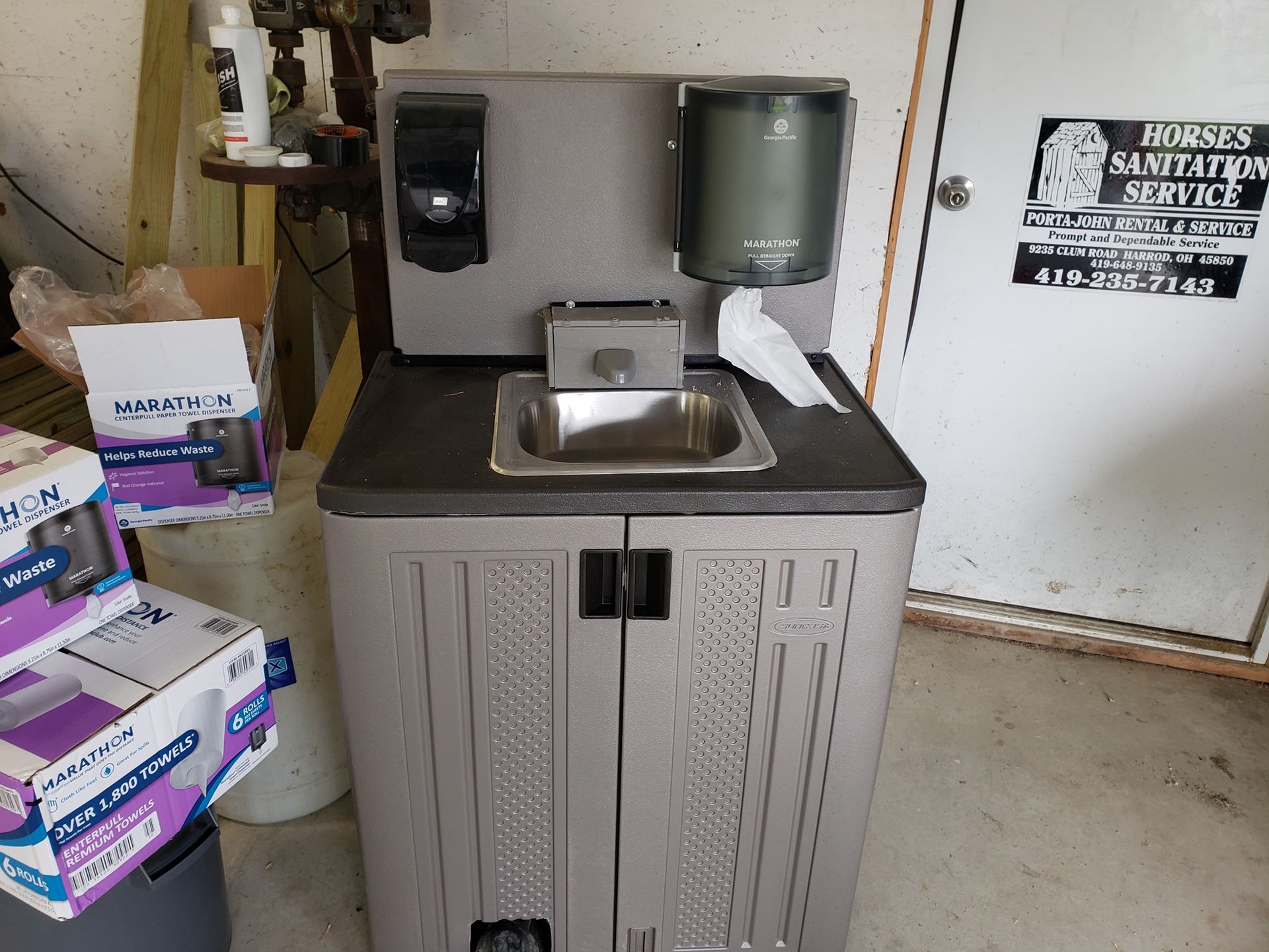 A grey, portable hand-washing station with a stainless steel sink, soap dispenser, paper towel holder, and foot pump.