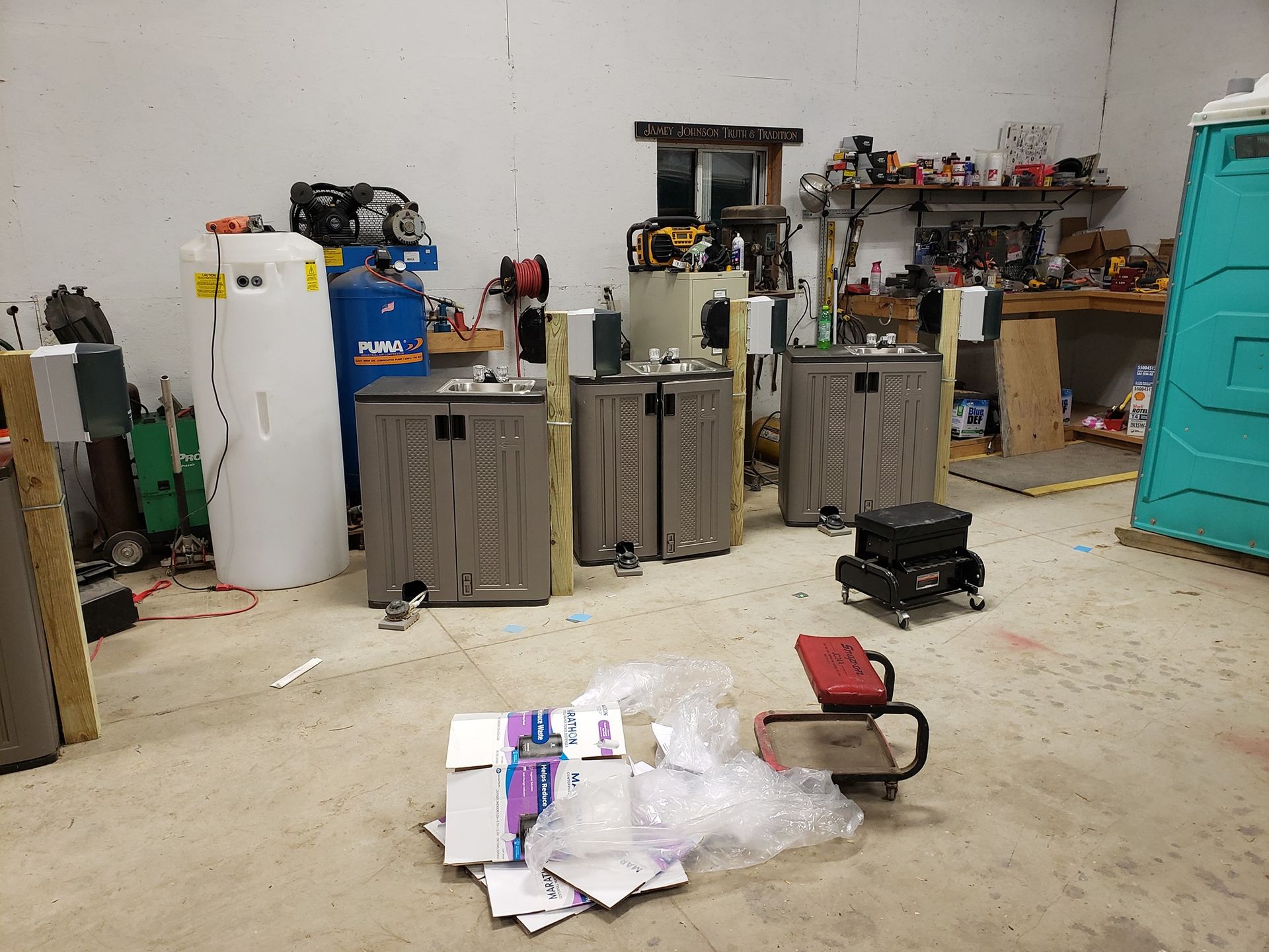 Workshop interior showing three small storage sheds, a white tank, a blue compressor, and scattered cardboard packaging.