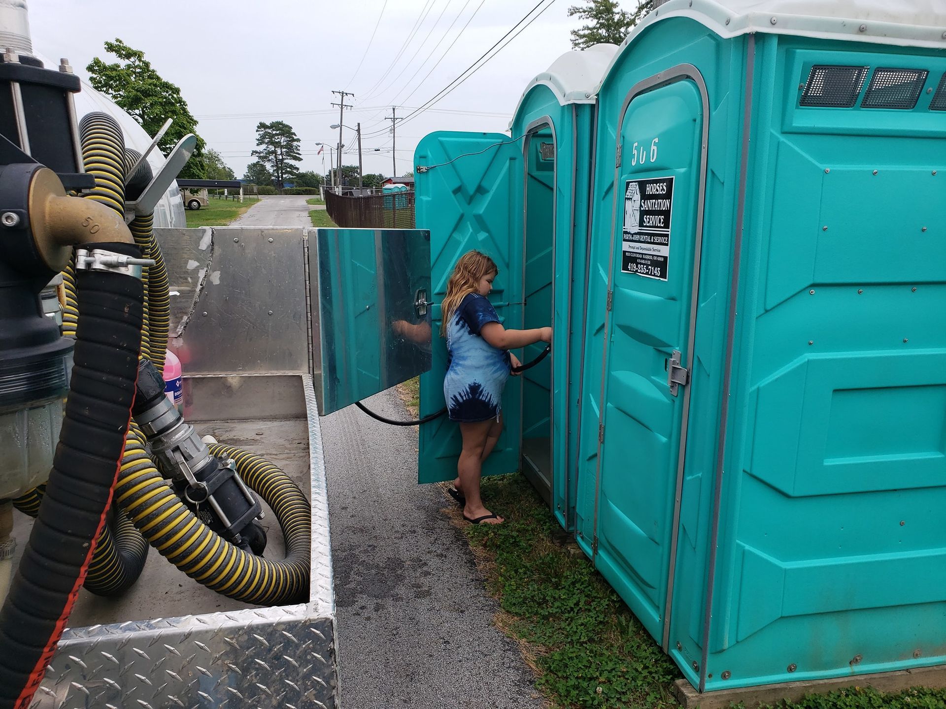 A worker services a bright teal portable toilet with a vacuum hose while standing outside in a gravel area.