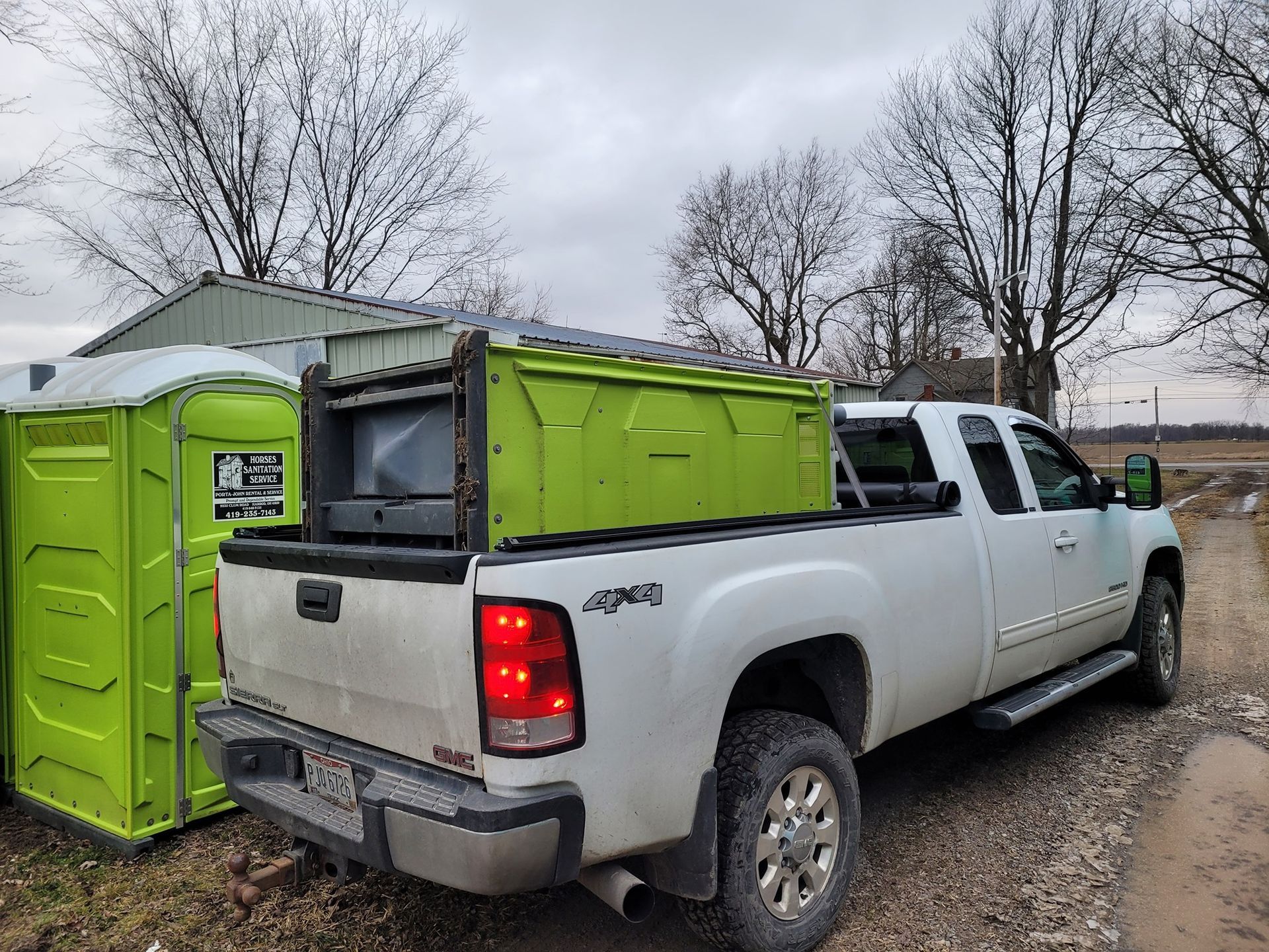 A white GMC pickup truck parked next to a green portable toilet, with a matching green storage container in its bed.