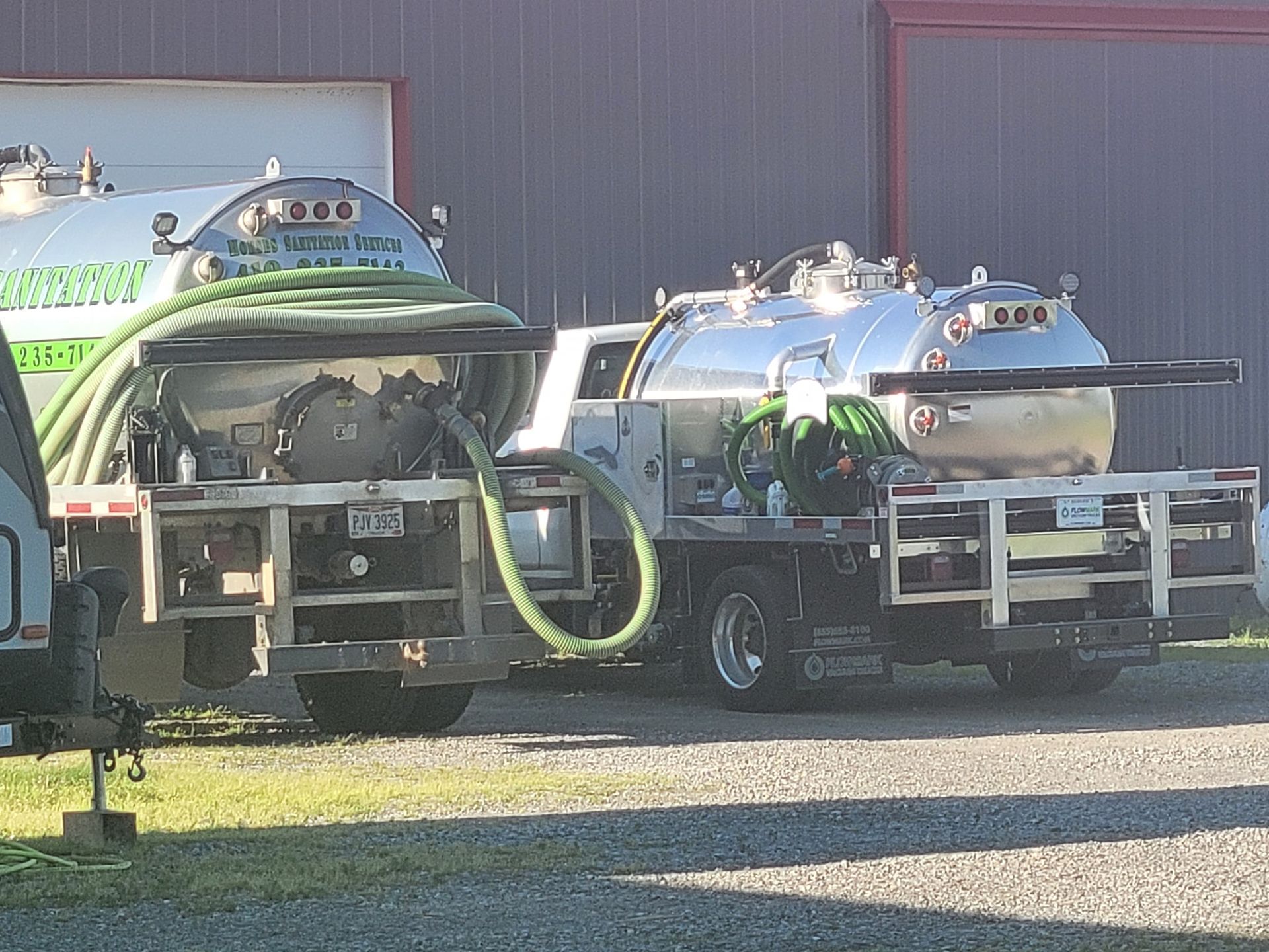 Two shiny, stainless steel tank trucks parked side-by-side on a gravel lot in front of a metal building.