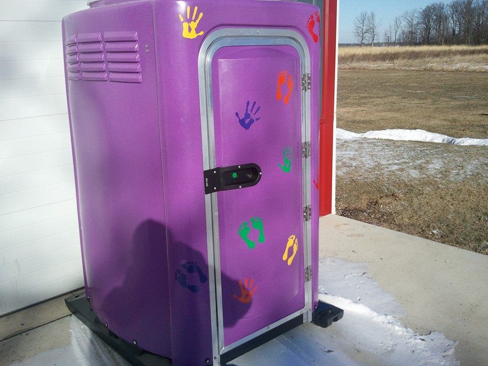 A purple portable restroom with colorful handprint and footprint decals on the door, sitting on a concrete surface.