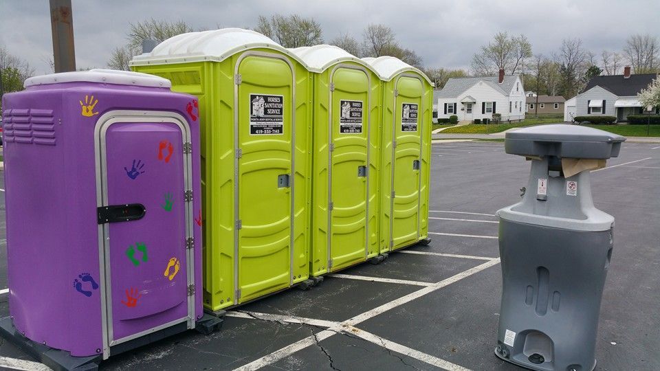 A row of one purple and three lime green portable toilets, with a portable handwashing station, parked on an asphalt lot.