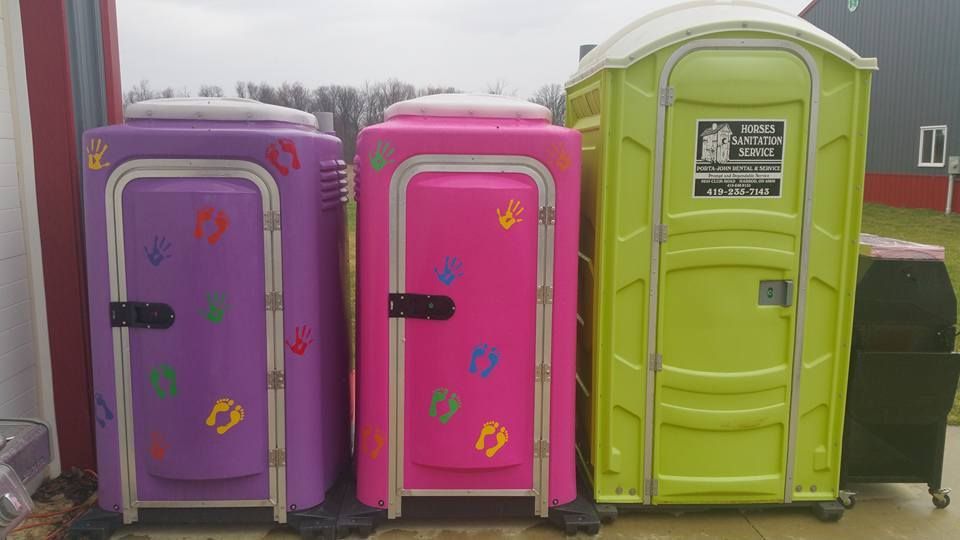 Three portable toilets standing in a row: one purple with colorful footprints, one pink, and one lime green.