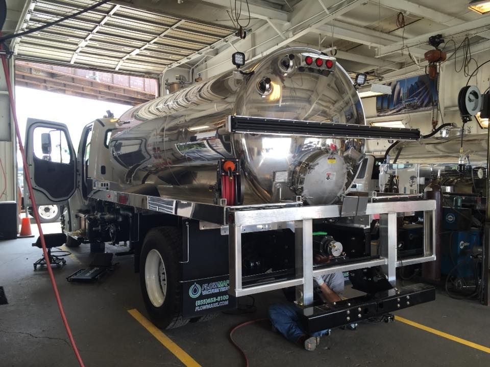 A chrome tanker truck parked in an indoor industrial garage, with a mechanic working underneath the rear frame.