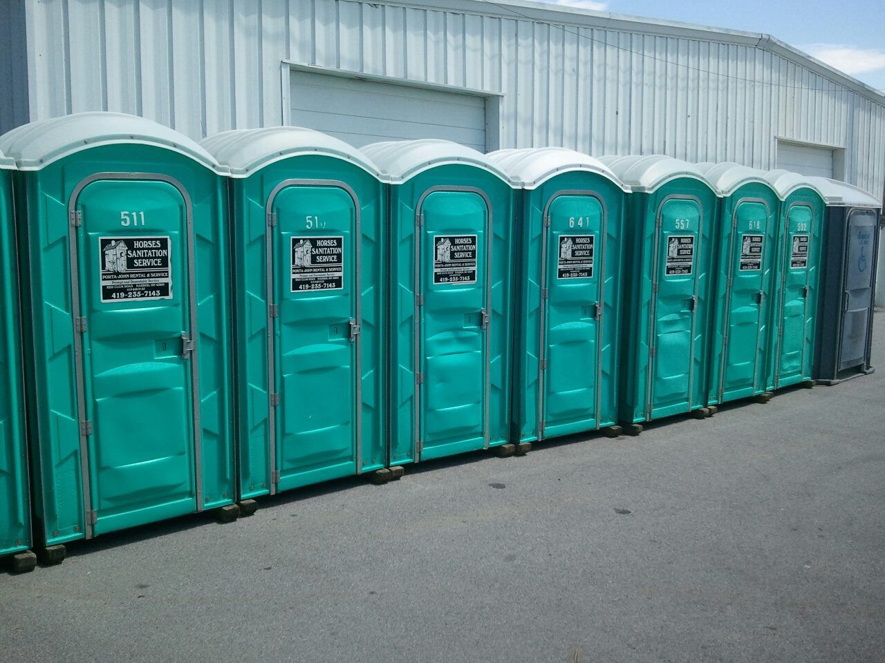 A row of teal portable toilets standing in a line on asphalt in front of a metal warehouse building.
