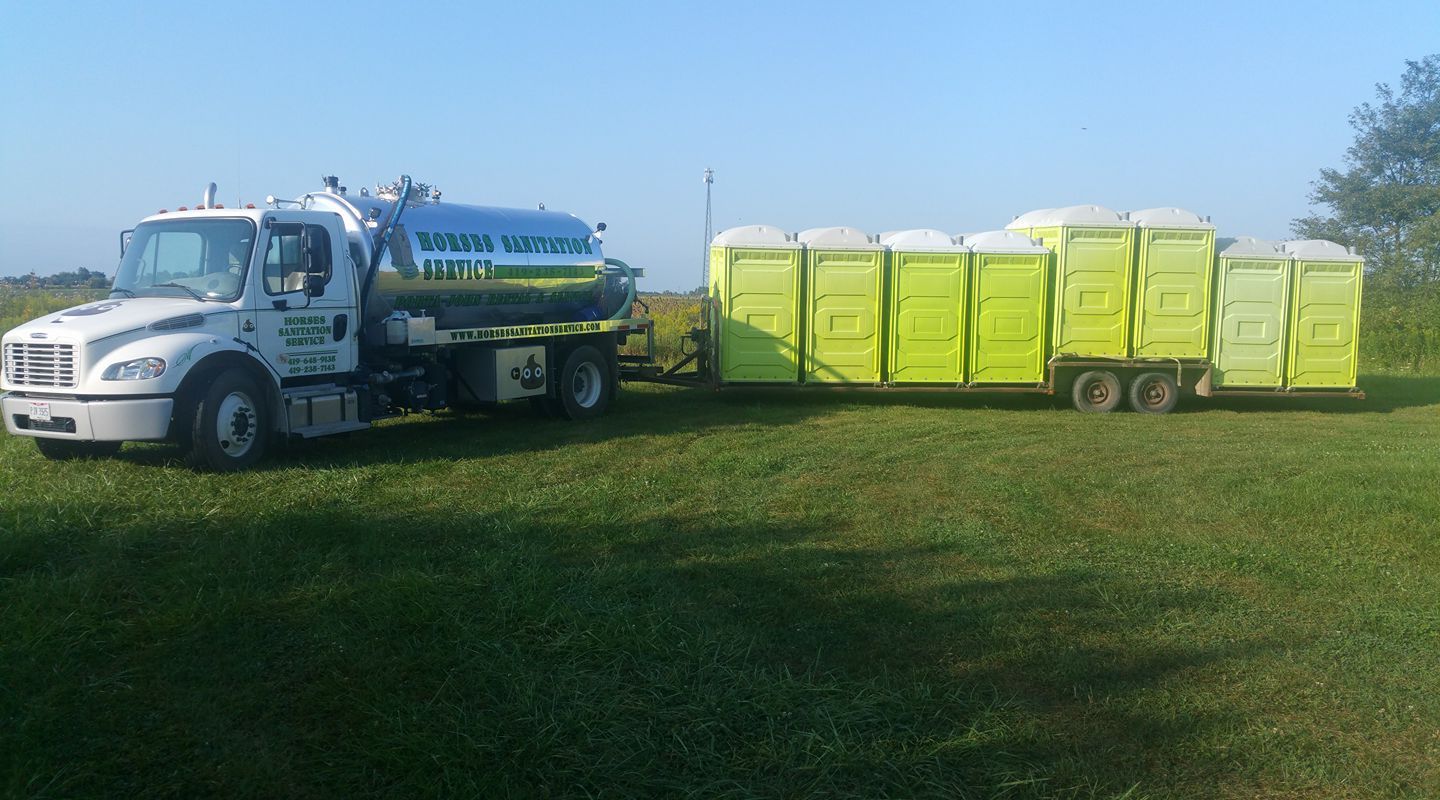 A white septic service truck is parked in a grassy field, pulling a trailer loaded with eight lime green portable toilets.