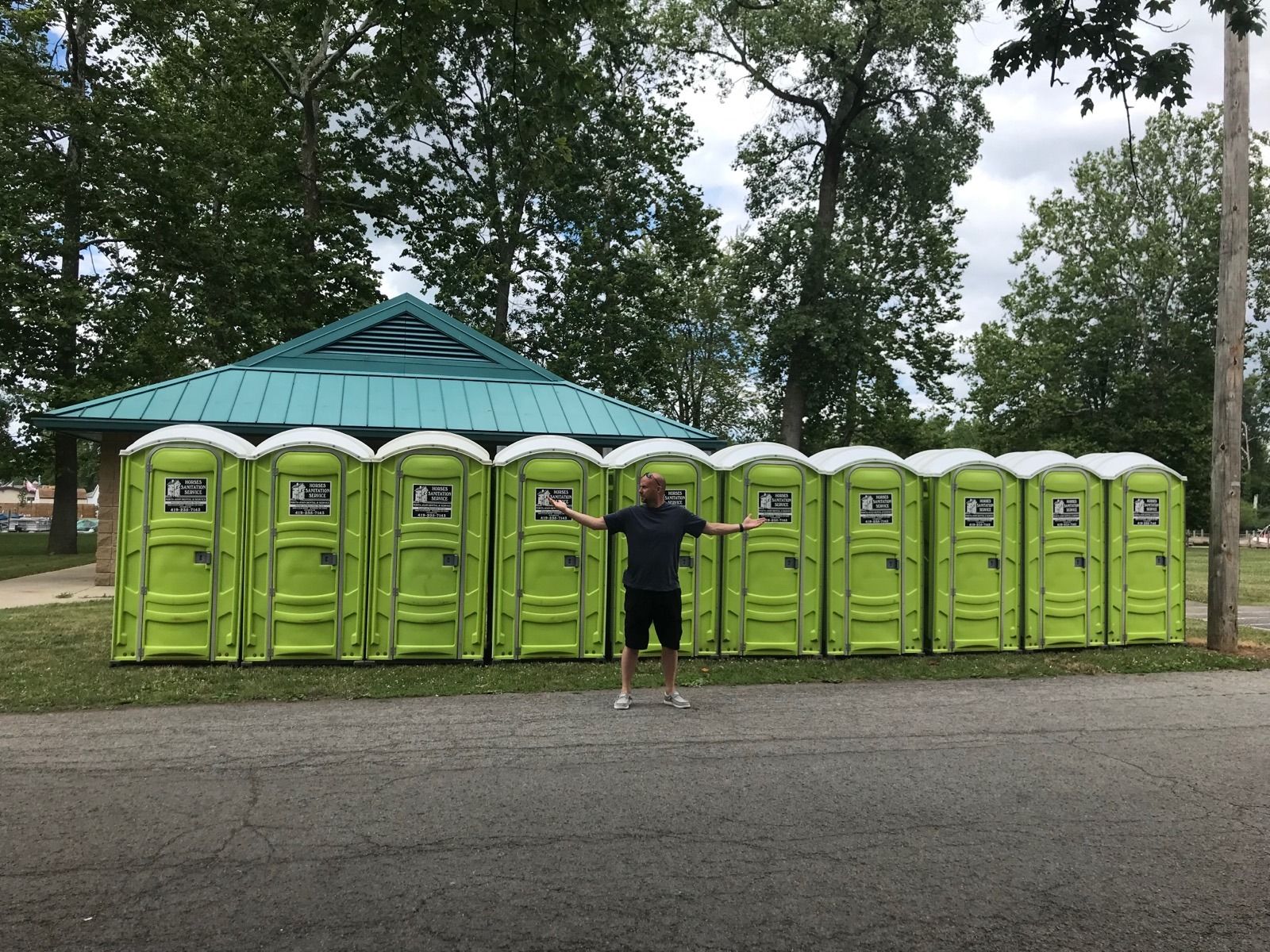 A person stands with arms spread wide in front of a long row of ten lime-green portable toilets near a park building.