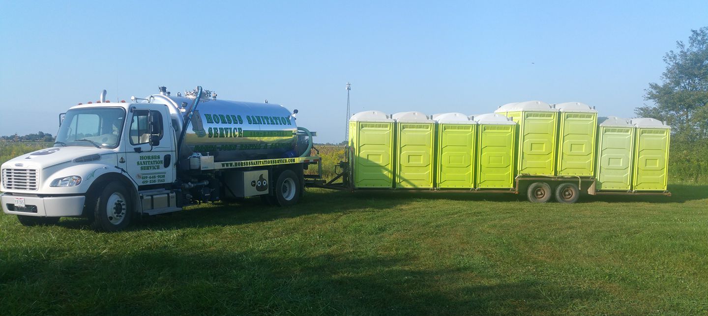 A white tanker truck towing a flatbed trailer loaded with several bright yellow portable toilets in a grassy field.