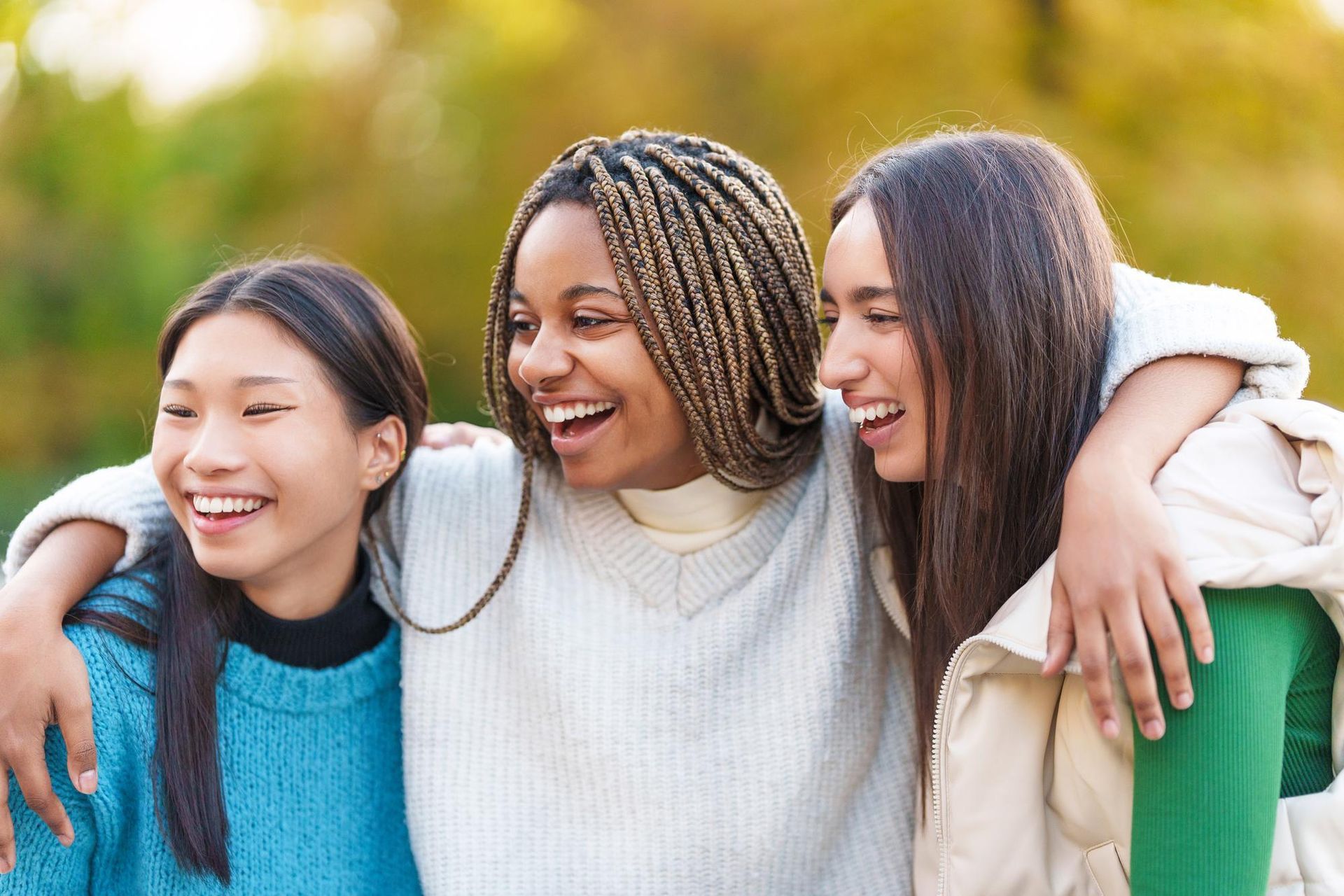 Three young women are standing next to each other in a park and smiling.
