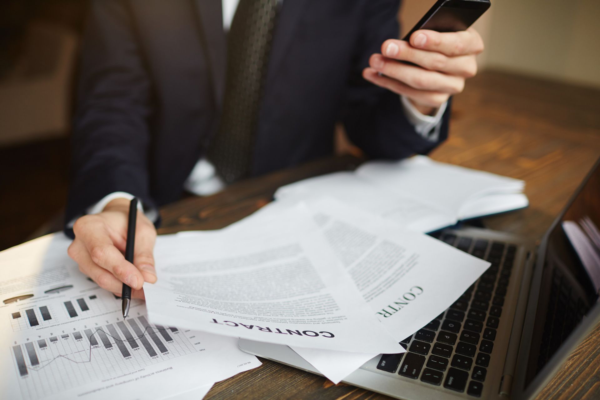 Person in a suit reviewing documents, holding a phone, and using a laptop. Papers and graphs are on the desk.