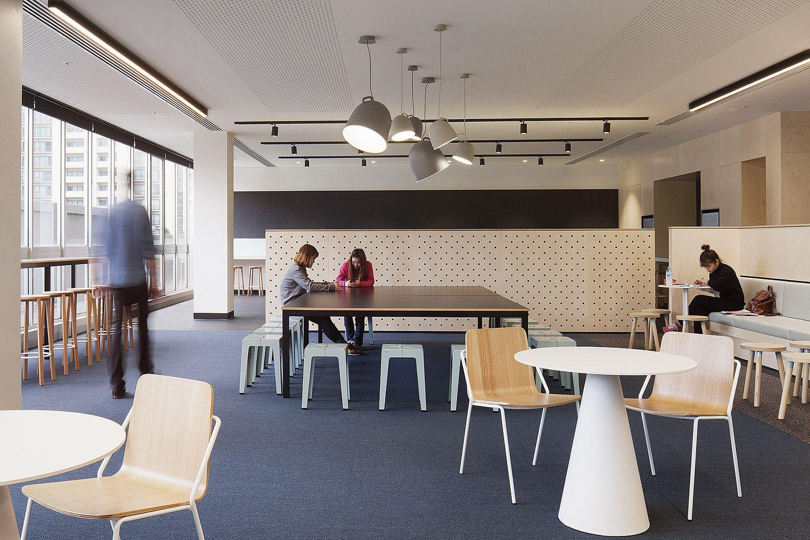 An office space with people working at tables. Blue carpet, white walls, and modern lighting.